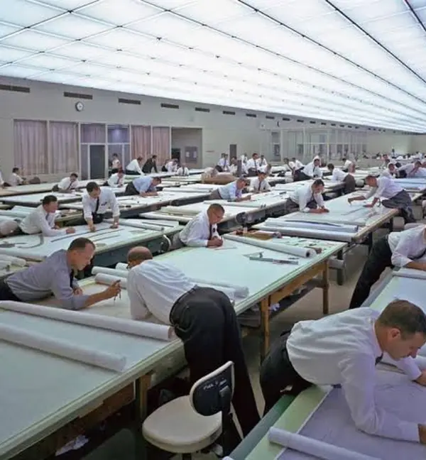 Dezenas de projetistas desenhando à mão em pranchetas, numa grande sala de desenho técnico